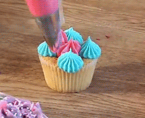 A close-up of a cupcake. Its top icing decoration is slowley being applied, one icing bag squeeze at a time with pink and blue icing.