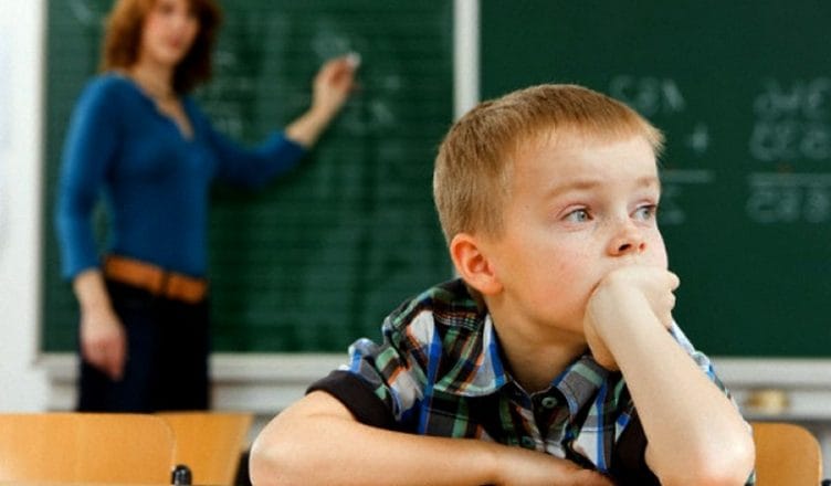 A young child looks off into the distance. A teacher stands at a blackboard behind them, looking for their attention.